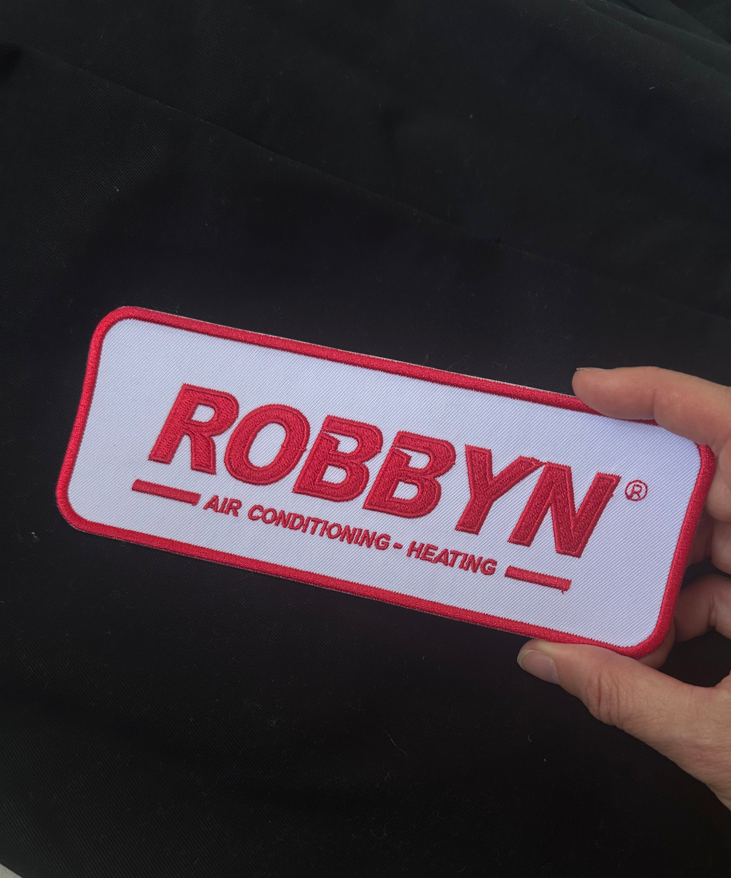 Hand holding a ROBBYN branded air conditioning and heating patch against a dark background