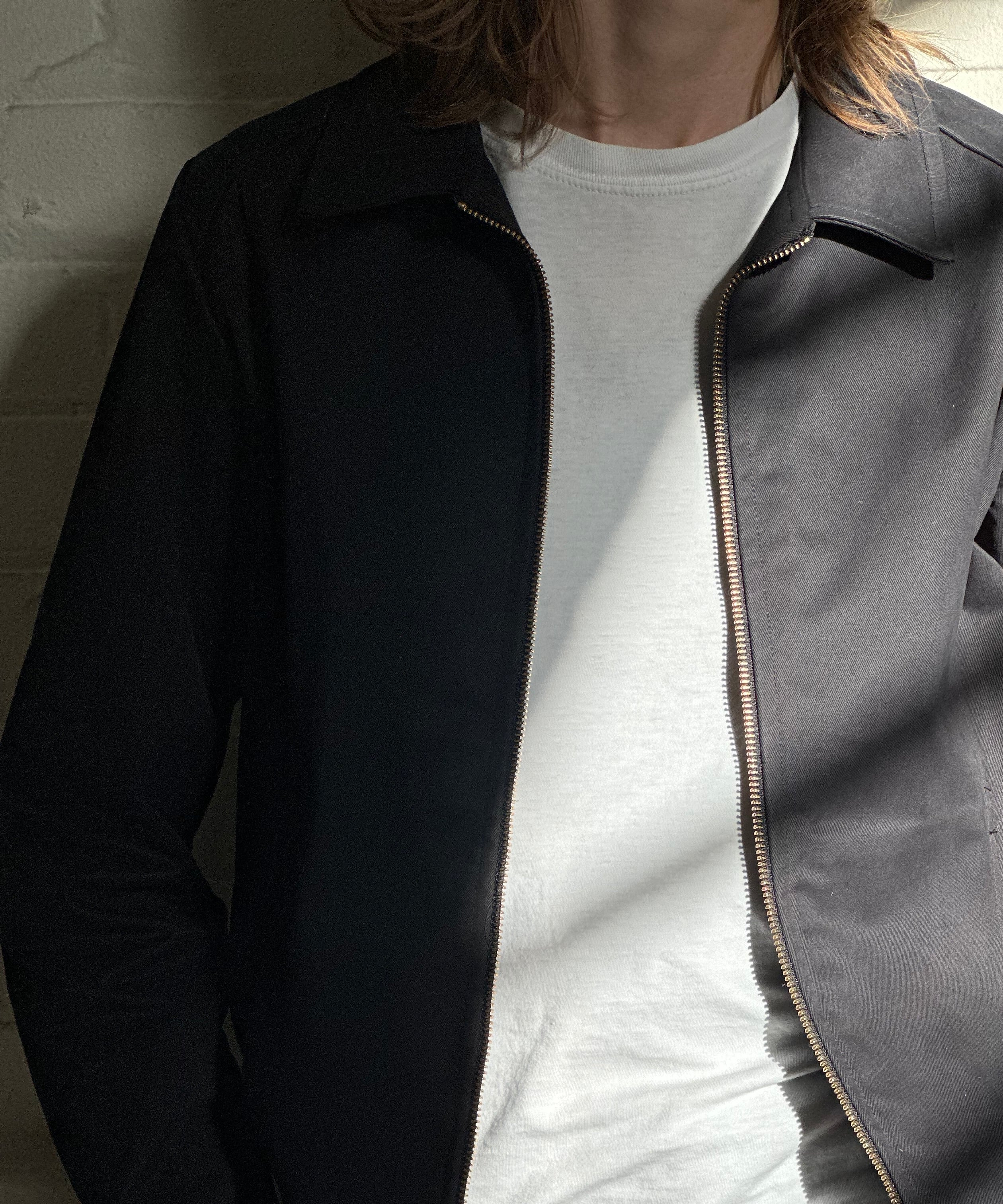 Close-up of the model wearing a black Mechano workwear jacket over a white T-shirt, standing in soft natural light against a white brick wall.