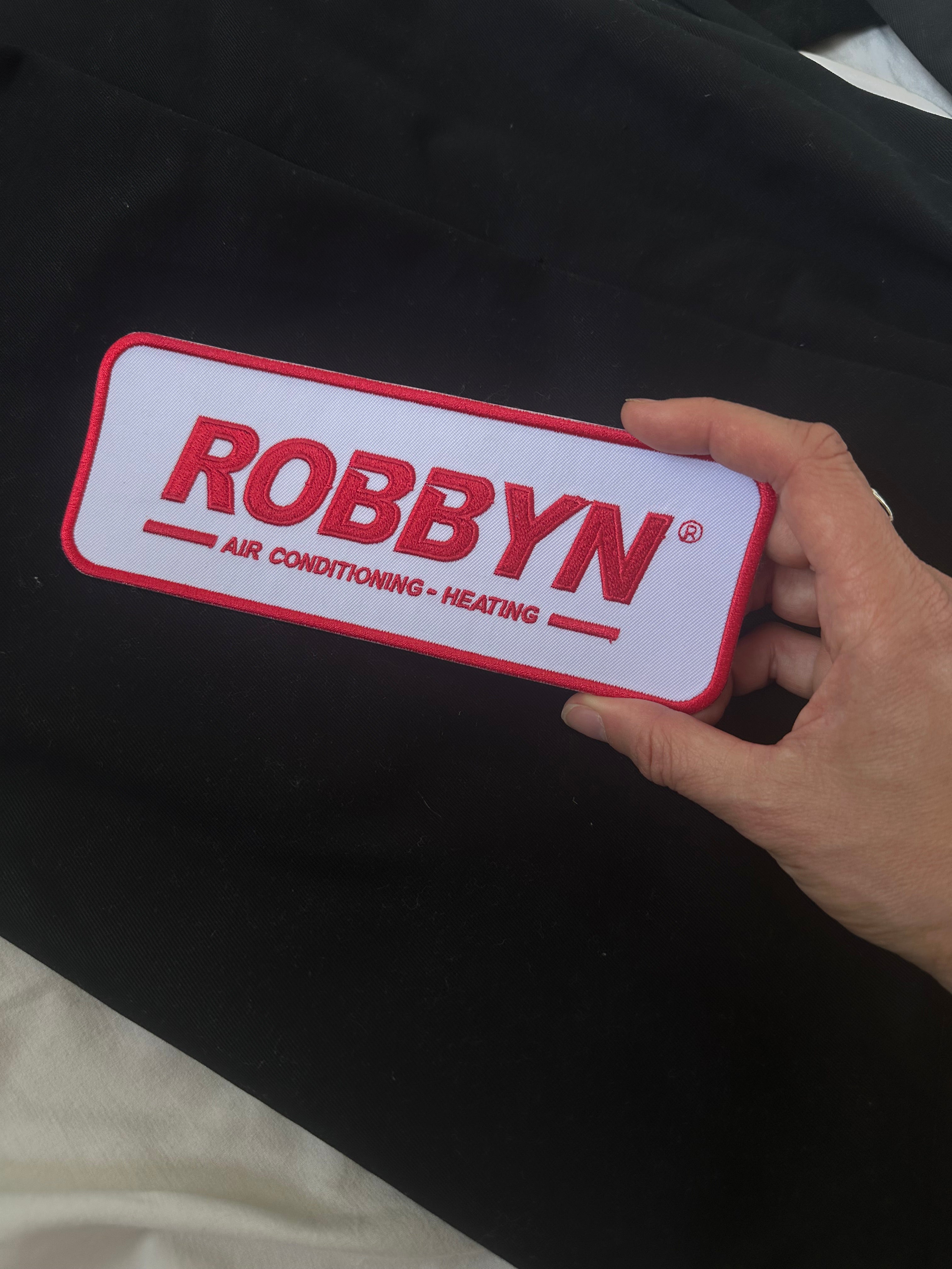 Hand holding a ROBBYN branded air conditioning and heating patch against a dark background