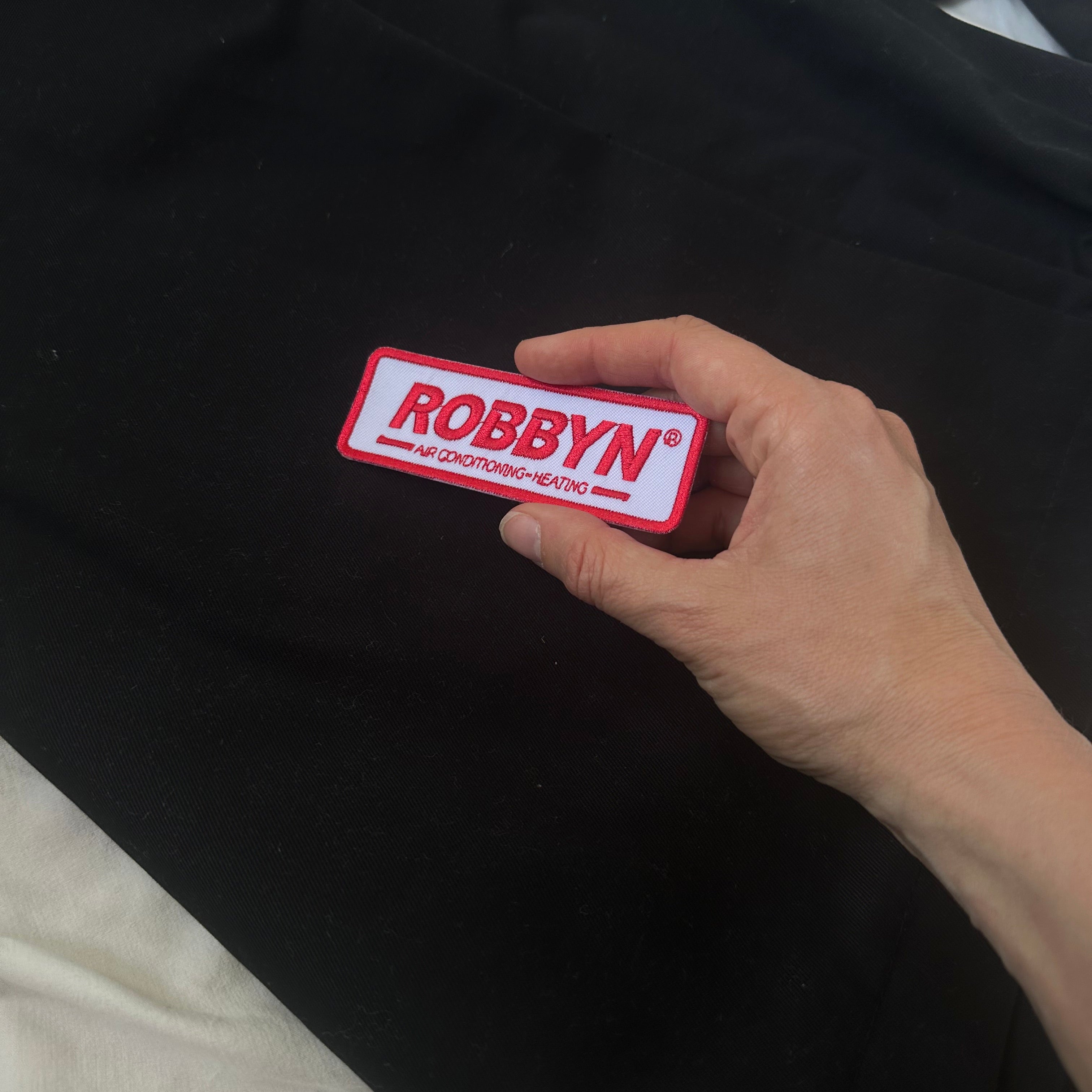 Hand holding a red and white 'ROBBYN' patch against a black background
