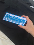 Hand holding a blue and white patch with 'Robbyn' 
