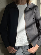 Close-up of the model wearing a black Mechano workwear jacket over a white T-shirt, standing in soft natural light against a white brick wall.